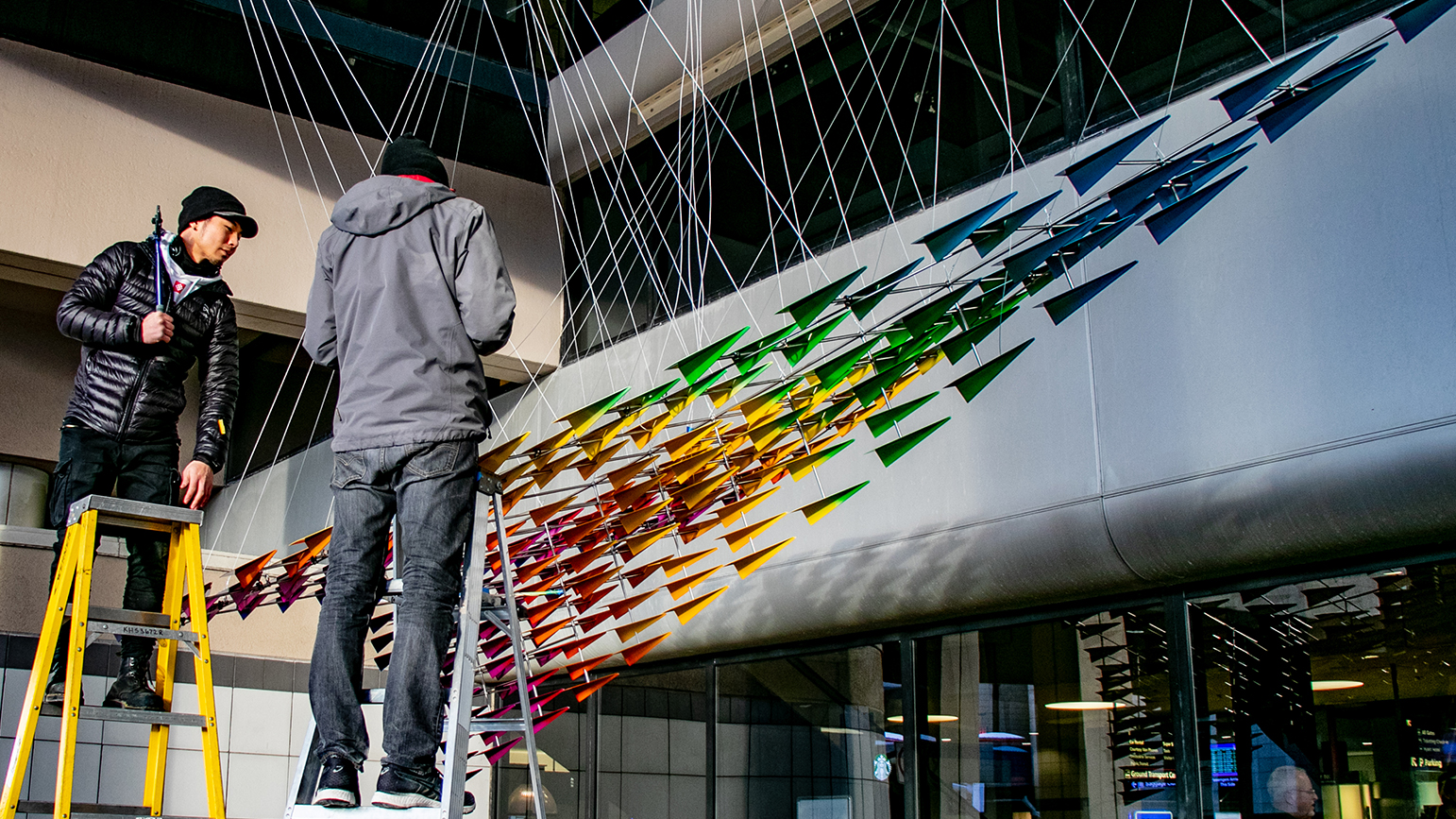 Image showing two people on ladders adding wiring to a large metal sculpture of rainbow-colored paper airplanes that together make up one large paper airplane