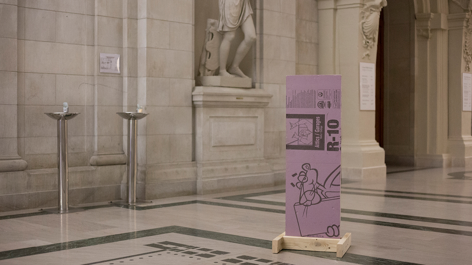 A photograph of a piece of installation on a simple wood stand in the middle of a beaux-arts hallway in the College of Fine Arts