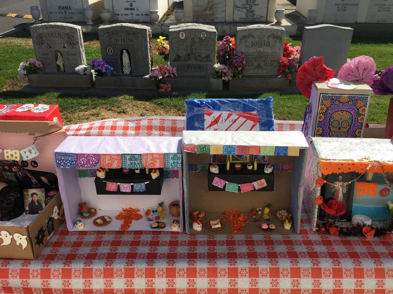 Mini ofrendas - shoeboxes filled with miniature foods, candy skulls, and other objects - sit on a checkered tablecloth in front of graves