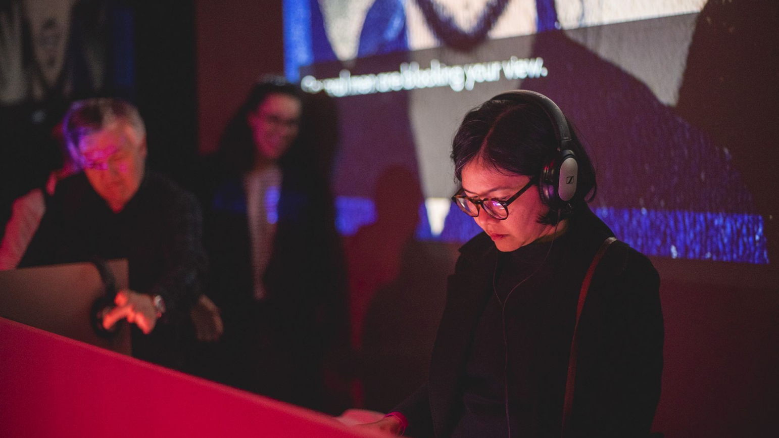 Photograph of people playing Angela Washko's "The Game: The Game" with a large screen behind them