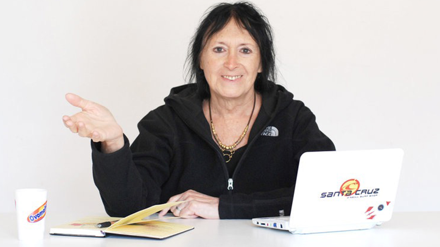 Photograph of Sandy Stone sitting at a desk with a notebook, laptop, and coffee cup