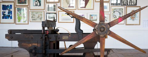 Photograph of a manual printing press with framed prints hung salon-style behind it.
