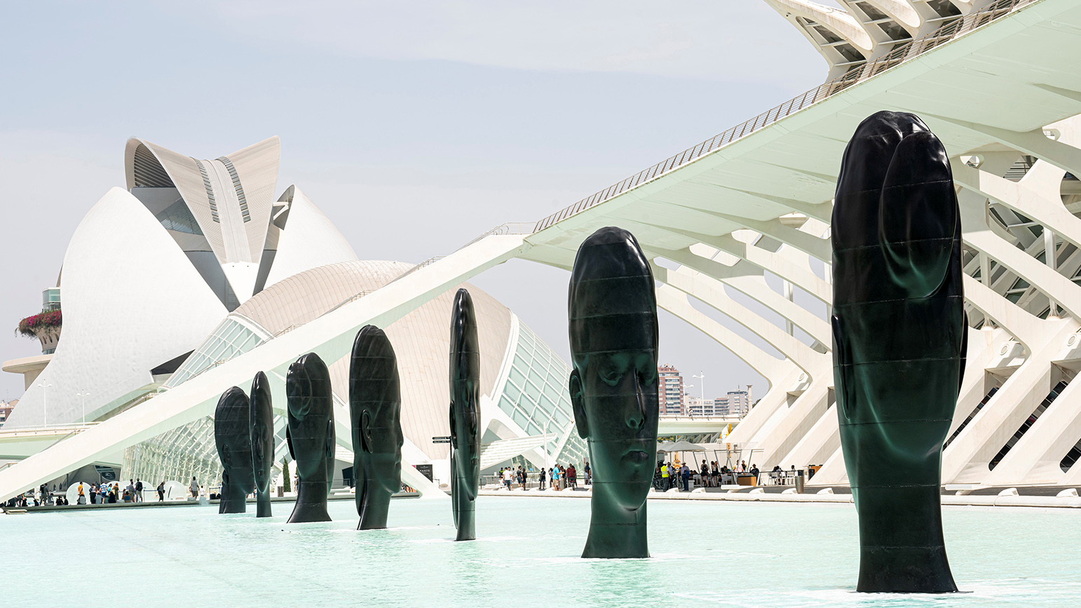 Seven large flat sculptures of serene faces installed in a shallow pool of water