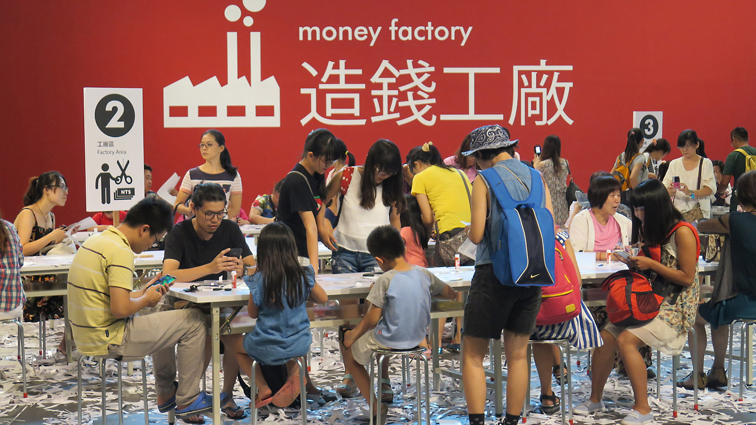 Large group of people sitting at tables with scraps of paper littering the floor and the words "money factory" on the wall behind them