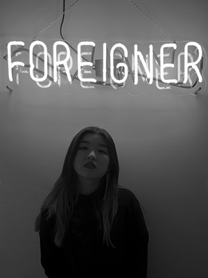 Photograph of Caroline Yoo with the word "FOREIGNER" behind her lit in neon