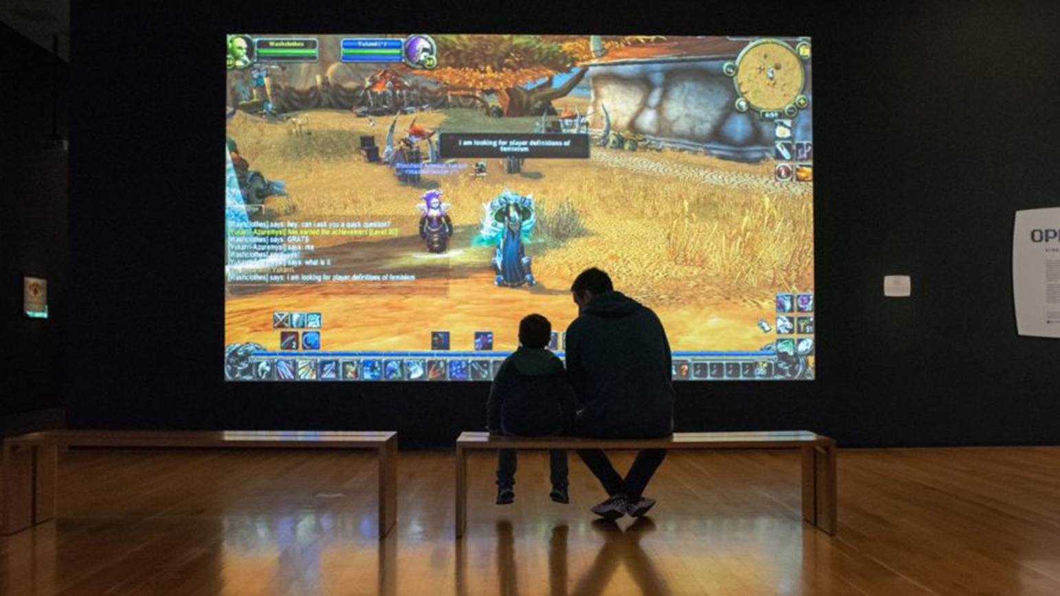 A child and adult sitting on a bench looking at a projection of the video game "World of Warcraft"