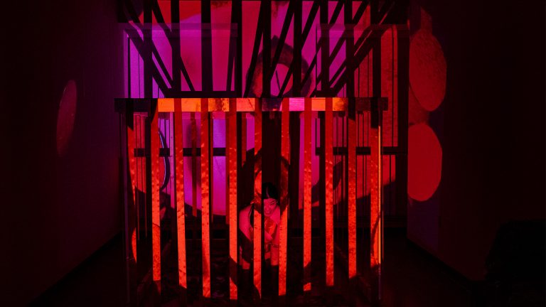 In a dark space with bright red and link light, a person is behind bars